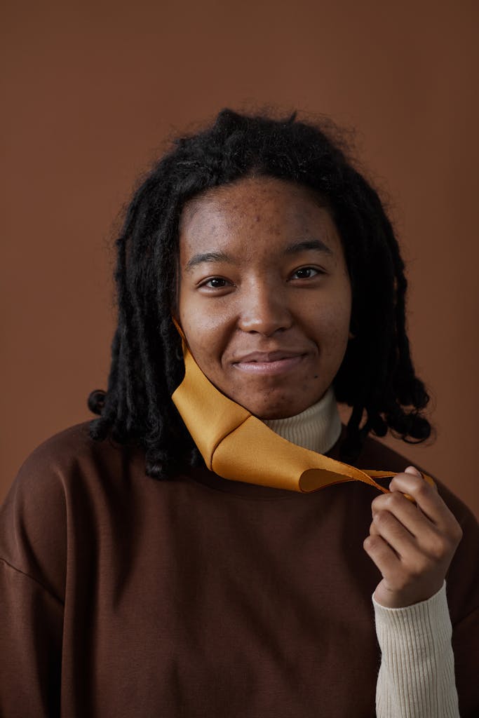 How To Start Accepting Your Skin And Feel More Confident A young woman with curly hair and acne removing a face mask while smiling indoors.