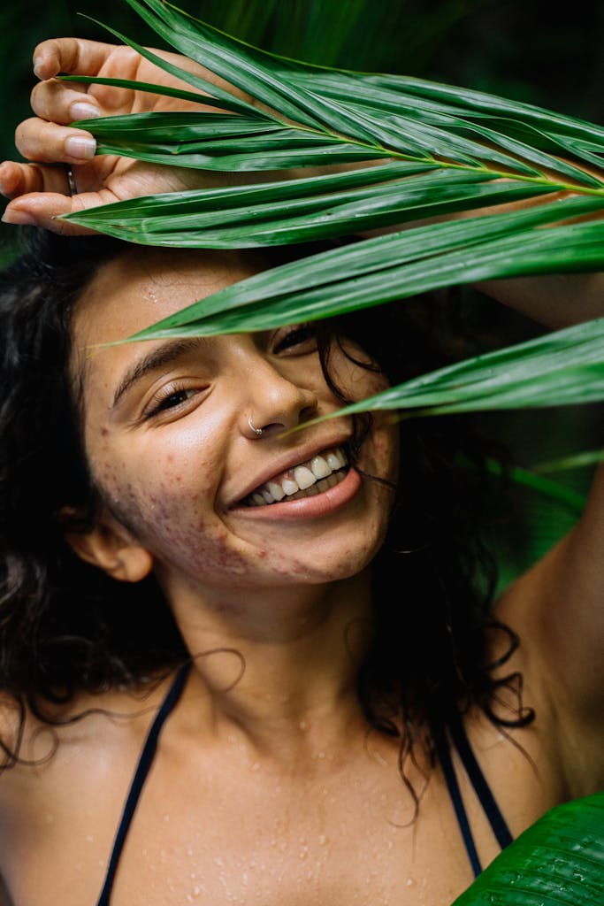 How To Start Accepting Your Skin And Feel More Confident Happy young woman with curly hair and nose piercing surrounded by lush green leaves.